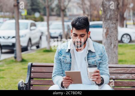 Un Indien à barbe, habillé avec élégance, est assis sur un banc et utilise sa tablette. Banque D'Images