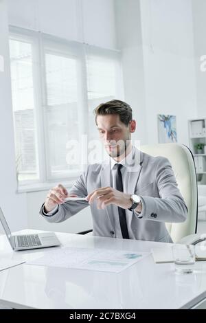 Jeune Manager élégant en costume gris assis au bureau et photographiant des graphiques sur un smartphone Banque D'Images