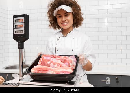 Foyer sélectif de jeune femme souriante avec des cheveux bouclés tenant la viande fraîche brute rouge dans les mains. Fille gaie derrière le comptoir du magasin montrant la viande dans le bol du réfrigérateur, supermarché. Banque D'Images