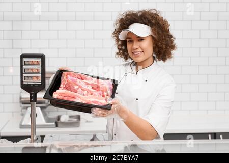 Foyer sélectif de jeune femme souriante avec des cheveux bouclés tenant la viande fraîche brute rouge dans les mains. Fille gaie derrière le comptoir du magasin montrant la viande dans le bol du réfrigérateur, supermarché. Banque D'Images