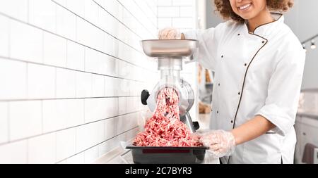 Incognito femme souriante avec des cheveux bouclés mettre de petits morceaux dans le moulin à viande et les grinds dans la viande hachée. Récolte de femmes travaillant dans la boucherie, préparant la viande pour la vente en magasin. Banque D'Images