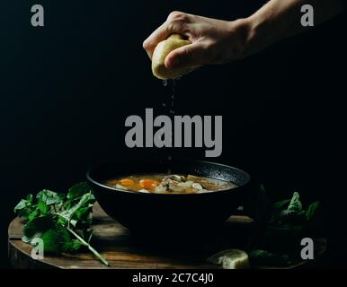 Personne pressant le citron sur la soupe dans un bol noir avec un arrière-plan sombre Banque D'Images