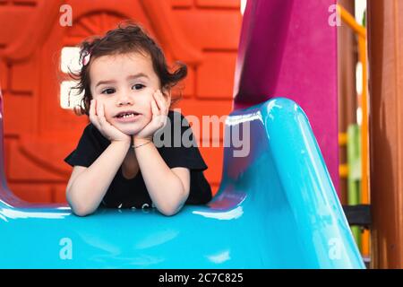 une mignonne fille de liitle qui pose sur la diapositive bleue dans un terrain de jeu avec les mains sous son menton. Banque D'Images