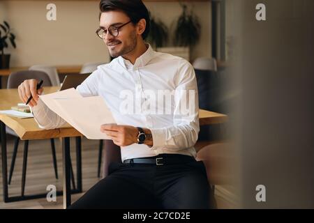 Jeune homme d'affaires beau dans les lunettes de vue heureusement travailler avec des papiers dans le bureau Banque D'Images