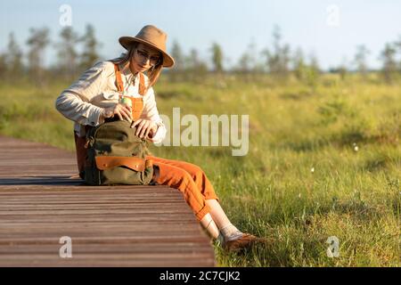 Jeune femme botaniste dans un chapeau assis sur un chemin en bois à travers le marais de tourbière dans le parc national de la faune.naturaliste reposant sur la promenade, prend un thermos f Banque D'Images