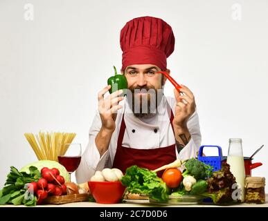 Faites cuire avec un visage heureux en uniforme bordeaux, assis près d'une table avec des légumes et des ustensiles de cuisine. Le chef prépare le repas. Un homme avec une barbe tient le piment et le poivron sur fond blanc. Concept de processus de cuisson Banque D'Images