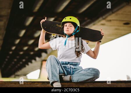 Gros plan de jeune fille attrayante avec planche à roulettes debout à l'extérieur dans le parc de skate. Une femme skate tenant son skate derrière. Skateboarder urbain Banque D'Images