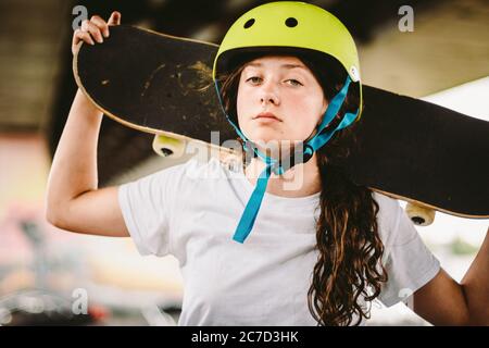 Gros plan de jeune fille attrayante avec planche à roulettes debout à l'extérieur dans le parc de skate. Une femme skate tenant son skate derrière. Skateboarder urbain Banque D'Images