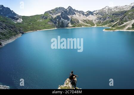 La silhouette de l'homme reste sur un pic de roche vif. Satisfaire les randonneurs profiter de la vue. Homme haut sur une falaise rocheuse regardant sur le lac bleu entouré de montagnes dans l'Al Banque D'Images