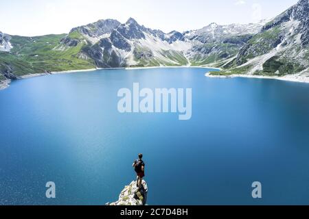 La silhouette de l'homme reste sur un pic de roche vif. Satisfaire les randonneurs profiter de la vue. Homme haut sur une falaise rocheuse regardant sur le lac bleu entouré de montagnes dans l'Al Banque D'Images