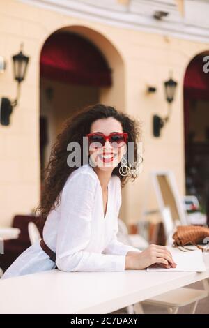 Dame gaie avec des cheveux mauriquement foncés dans des lunettes de soleil debout et regardant joyeusement dans l'appareil-photo tout en passant du temps en terrasse d'été du café Banque D'Images