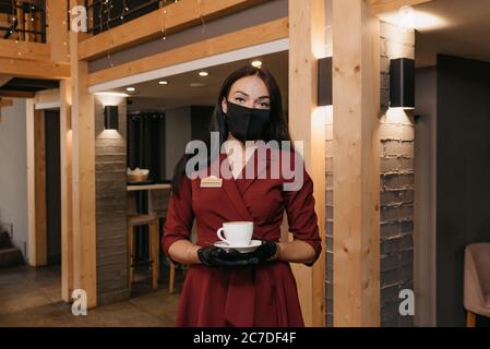 Une magnifique directrice de restaurant qui porte un masque noir et des gants jetables tient une tasse de café dans un restaurant. Un propriétaire de café aimable Banque D'Images