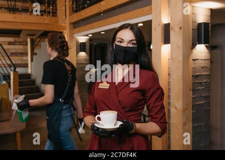 Une magnifique directrice de restaurant qui porte un masque noir et des gants jetables tient une tasse de café dans un restaurant. Une serveuse est propre Banque D'Images