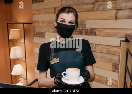 Une jolie serveuse qui porte un masque noir et des gants jetables sert une tasse de café dans un restaurant. Un barista mignon attend les clients Banque D'Images