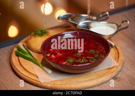 Photo d'un borsch ukrainien avec un pampuchka, un oignon vert et une crème sure sur un plateau en bois sur la table d'un restaurant. Banque D'Images