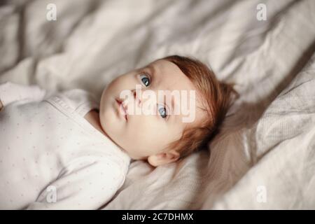 Portrait en gros plan de bébé casien mignon nouveau-né. Adorable bébé drôle avec des yeux gris bleu et des cheveux rouges allongé sur le lit et regardant loin. Authentique Banque D'Images