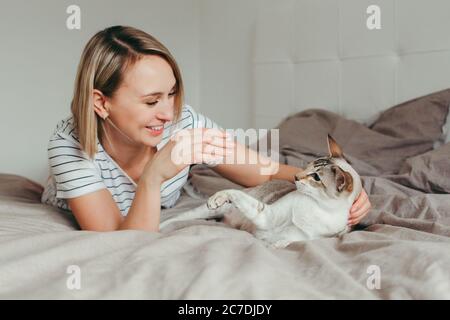 Portrait d'une femme blonde souriante et heureuse de race blanche couchée sur un lit dans la chambre à la maison et jouant avec un chat oriental de couleur point. Propriétaire de l'animal Banque D'Images