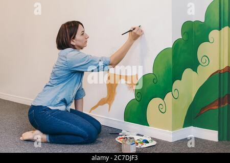 Femme caucasienne artiste peinture à la main peintures murales sur les murs à l'intérieur de l'appartement ou de l'école de studio avec des peintures acryliques. Mode de vie créatif passe-temps et freelance Banque D'Images