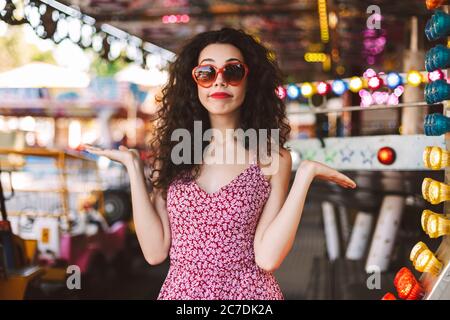 Jeune femme avec des cheveux en robe maurique foncé debout et montrant les mains sur le côté tout en regardant avec soin dans l'appareil photo dans le parc d'attractions Banque D'Images