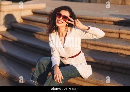 Une dame joyeuse avec des cheveux en forme de curly foncé dans des lunettes de soleil et une veste blanche assise sur les escaliers de la rue et regardant avec joie dans l'appareil photo tout en montrant deux doigts Banque D'Images