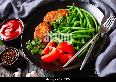 gros plan sur des viandes hachées en polenta, servies avec des haricots verts blanchis et des tomates fraîches sur une assiette noire sur une table en bois sombre, horizontale v Banque D'Images