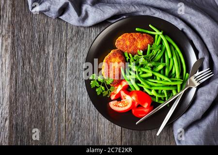 viandes de poulet émincées en croûte de maïs servies avec des haricots verts cuits à la vapeur et des tomates fraîches sur une assiette noire sur une table en bois sombre, vue horizontale fr Banque D'Images