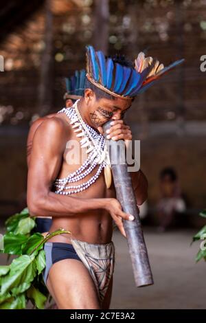 Un indigène amazonien portant une coiffe à plumes et de la peinture faciale joue un instrument cérémoniel, célébrant les traditions culturelles du Brésil. Banque D'Images