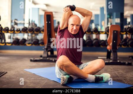 Un portrait d'homme âgé chauve qui s'étire après l'entraînement dans la salle de gym. Concept de personnes, de soins de santé et de mode de vie Banque D'Images