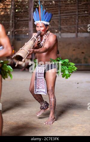 Un indigène amazonien portant une coiffe à plumes et de la peinture faciale joue un instrument cérémoniel, célébrant les traditions culturelles du Brésil. Banque D'Images