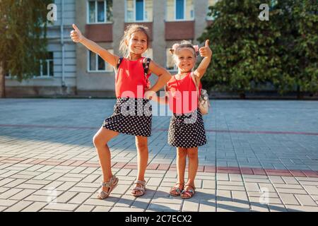 Les filles joyeuses sœurs portent des sacs à dos et montrent les pouces. Enfants élèves ayant du plaisir à l'extérieur bâtiment d'école. Banque D'Images