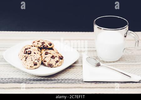 Gros plan de biscuits aux pépites de chocolat dans une assiette blanche à côté d'une tasse en verre transparent remplie de lait Banque D'Images