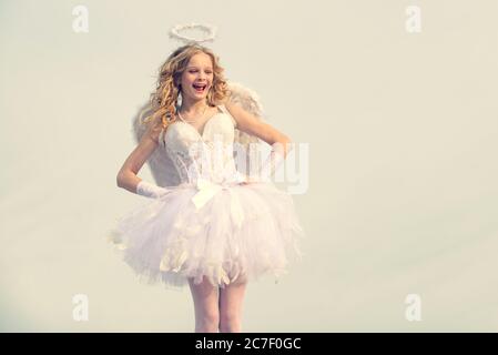 Innocent fille avec ailes d'ange debout avec arc et flèche contre ciel bleu et nuages blancs. Portrait d'une petite fille de cupidon priez. Enfant avec angélique Banque D'Images