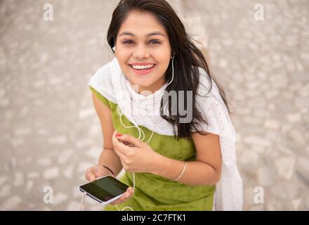 Belle jeune fille indienne heureuse écoutant de la musique sur smartphone à travers des écouteurs en plein air et regardant l'appareil photo avec un sourire éclatant. Banque D'Images