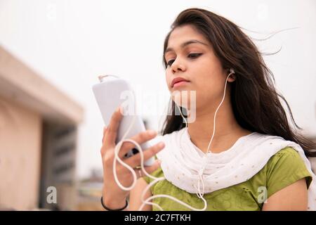 Belle et sereine jeune fille indienne tardive écoutant de la musique sur smartphone à travers des écouteurs et en profitant de l'air frais en extérieur sur le toit à l'heure de la journée. Banque D'Images
