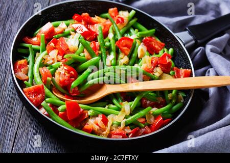 Gros plan de la poêle haricots verts frits avec oignons et tomates, faire frire le bodi dans une poêle sur une table en bois avec un chiffon gris, vue horizontale depuis le dessus Banque D'Images