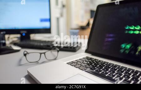 Vue rapprochée et peu profonde d'un ordinateur portable bien connu sur un bureau, avec un moniteur connecté à un serveur et une paire de lunettes. Banque D'Images