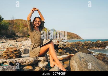 femme blogueur travaillant à distance avec un ordinateur portable utilisant l'internet, sur la mer, assis sur la plage. Tient un ordinateur portable sur ses genoux et regarde à travers Banque D'Images