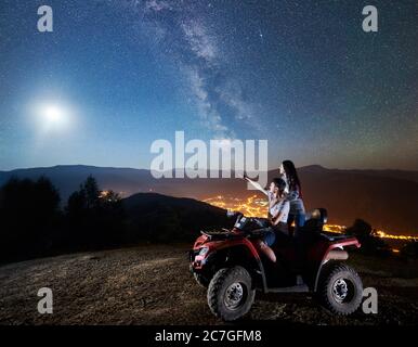 Jeune couple homme et femme touristes à cheval vtt quad moto sur le sommet de la montagne. Homme pointant vers un beau ciel nocturne plein d'étoiles, pleine lune, voie lactée, village lumineux sur fond Banque D'Images