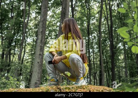 Jeune fille avec de longs cheveux assis sur une bûche d'arbre dans la forêt de printemps. Femme randonneur se reposant Banque D'Images