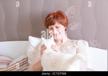 Femme âgée agréable avec des cheveux rouges courts dans les vêtements de maison est assis sur le lit et petting un chat blanc mignon. Vie lente. Copier l'espace Banque D'Images