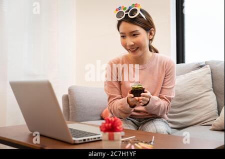 Femme asiatique célébrant son anniversaire par vidéoconférence en ligne, à la maison, tout en restant isolée à la maison. Main tenant un gâteau de tasse Banque D'Images