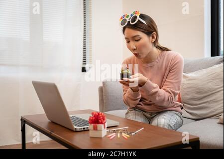 Femme asiatique célébrant son anniversaire par vidéoconférence en ligne, à la maison, tout en restant isolée à la maison. Main tenant un gâteau de tasse Banque D'Images