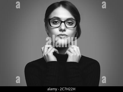 Portrait d'une jeune fille étudiante portant un pull à col roulé noir et de lunettes stylées posé sur fond gris en studio Banque D'Images