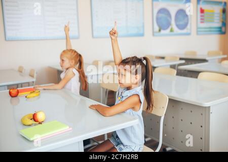 Deux petites écolières s'assoient à un bureau dans une classe scolaire et lèvent leurs mains Banque D'Images