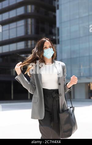 Une jeune femme entreprenante se promenant résolument entre les immeubles de bureaux. Elle porte un masque médical comme mesure préventive pour la crise de santé Covid-19. Banque D'Images