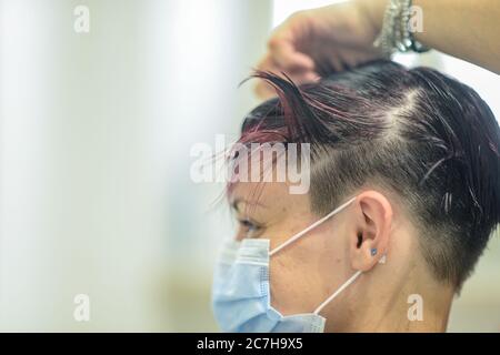 Jeune femme attirante dans un salon de beauté coupant le style et rouge mourir par un styliste professionnel portant un masque de protection contre la contagion Banque D'Images