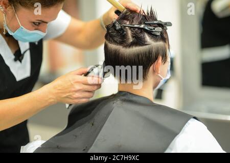 Jeune femme attirante dans un salon de beauté coupant le style et rouge mourir par un styliste professionnel portant un masque de protection contre la contagion Banque D'Images