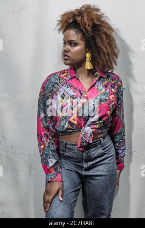 Jeune femme afro-américaine tendance avec coiffure afro et boucles d'oreilles à pampilles jaunes portant une chemise tendance colorée avec noeud et un Jean élégant sta Banque D'Images