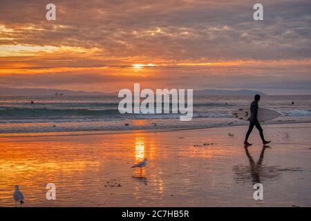 Un garçon avec une planche de surf à pied à travers la plage entourée la mer ondulée pendant le coucher de soleil doré Banque D'Images