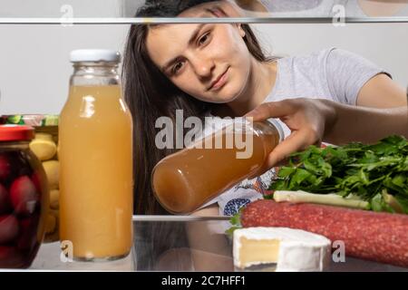 La fille regarde dans le réfrigérateur et prend le jus de fruit dans une bouteille de verre. Produits dans le réfrigérateur. Banque D'Images